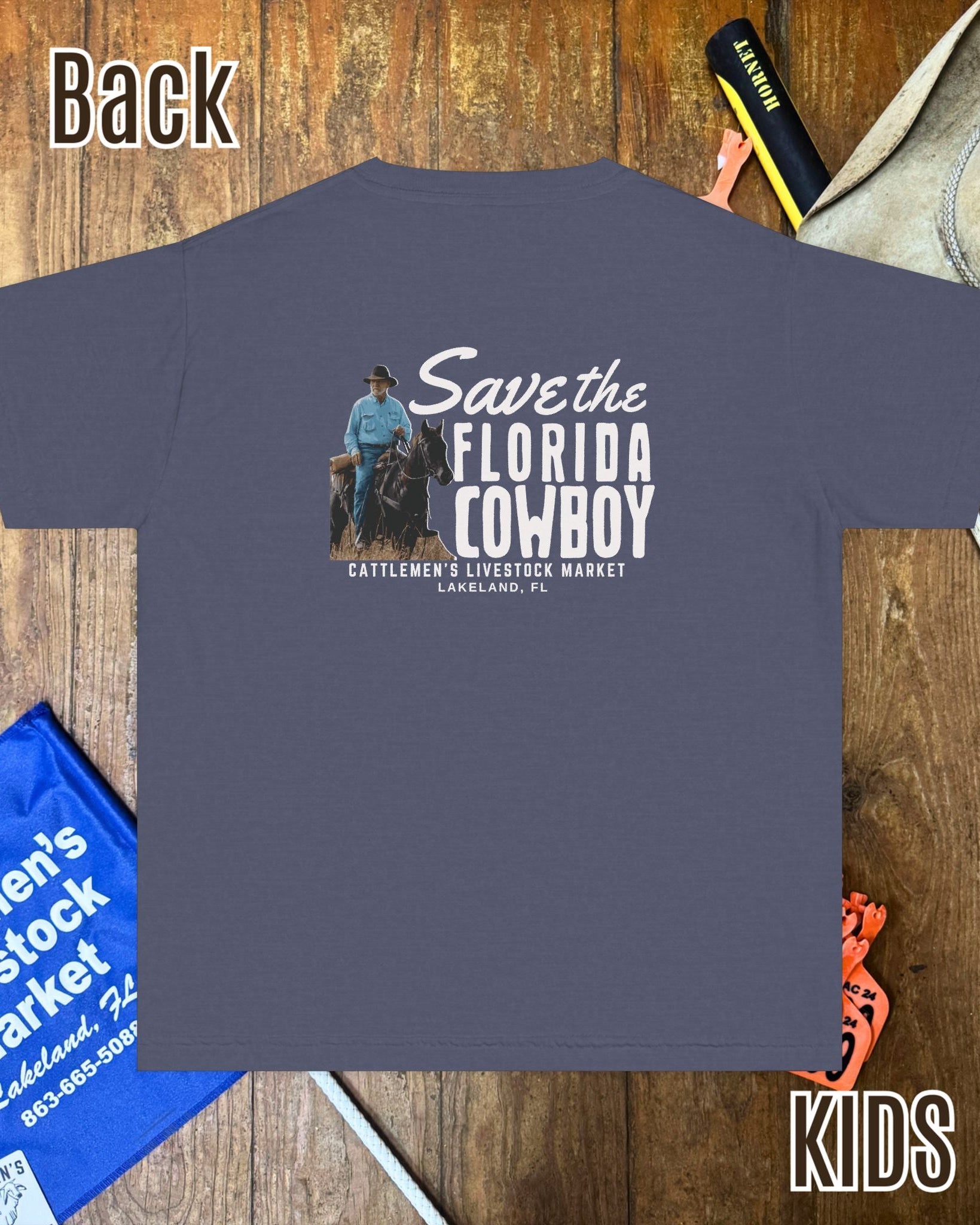 A dark slate blue kids' t-shirt is displayed flat on a rustic wooden background with western-themed props like a cowboy hat, a blue market bag, and a yellow marker. The back of the shirt features a graphic of a cowboy on a tractor with the text 'Save the Florida COWBOY'.