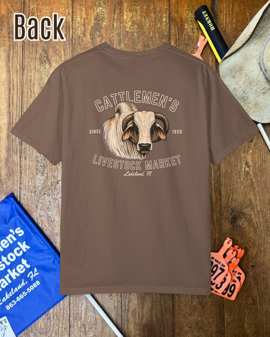 A brown short-sleeve T-shirt shown from the back, laid flat on a wooden surface with western-themed props including a cowboy hat, a blue sign, and an orange ticket. The shirt features a large graphic of a ram's head with the text 'CATTLEMEN'S LIVESTOCK MARKET' and 'Leland, TX'.