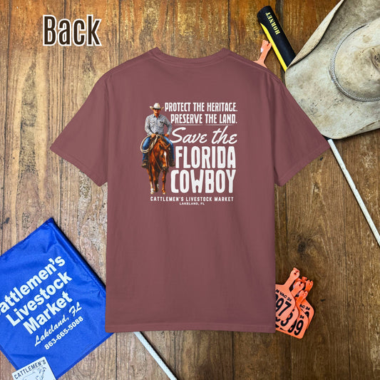 A mauve-brown short-sleeve graphic T-shirt laid flat on a wooden surface, surrounded by cowboy-themed props including a hat, boots, a blue market bag, and an orange ticket. The shirt features a large western-style graphic with a cowboy on a horse and the text 'Protect the Heritage Preserve the Land.