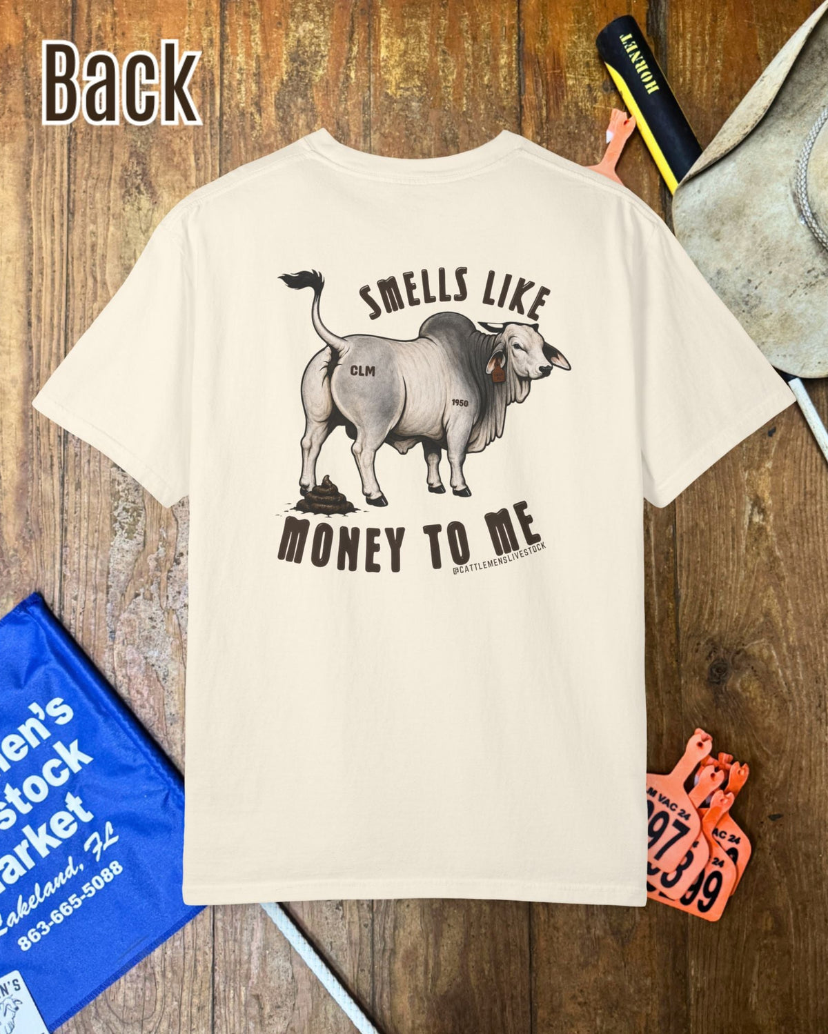 A cream-colored short-sleeve t-shirt with a western-themed graphic on the back, featuring a steer and the text 'SMELLS LIKE MONEY TO ME'. The shirt is displayed in a flat-lay arrangement on a rustic wooden background alongside a cowboy hat, a blue leather bag, and an orange ticket.