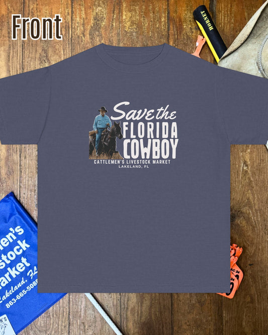 A dark slate blue graphic t-shirt laid flat on a rustic wooden background, surrounded by cowboy-themed props including a hat, boots, and a blue sign. The t-shirt features a graphic with a cowboy on a horse and the text 'Save the FLORIDA COWBOY' and 'Cattlemen 2 Livestock Market LADYSON, FL'.