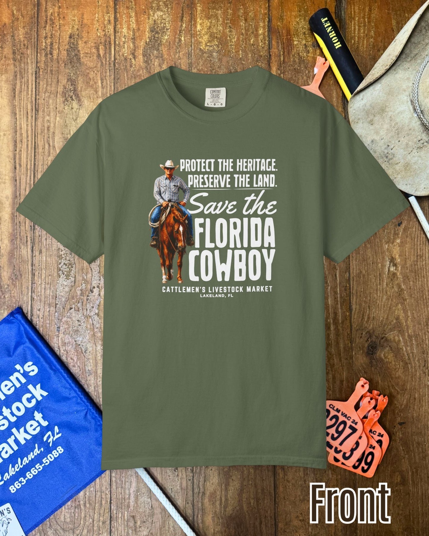 An olive green graphic t-shirt laid flat on a rustic wooden background, surrounded by cowboy-themed props including a hat, a blue sign, and an orange ticket. The t-shirt features a graphic of a cowboy on a horse with the text 'Protect the heritage.