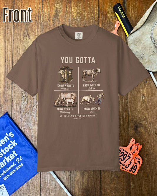 A brown graphic t-shirt featuring a western-themed cattle market design, displayed in a flat-lay arrangement on a wooden background with a cowboy hat, a blue tote bag, and cattle tags.