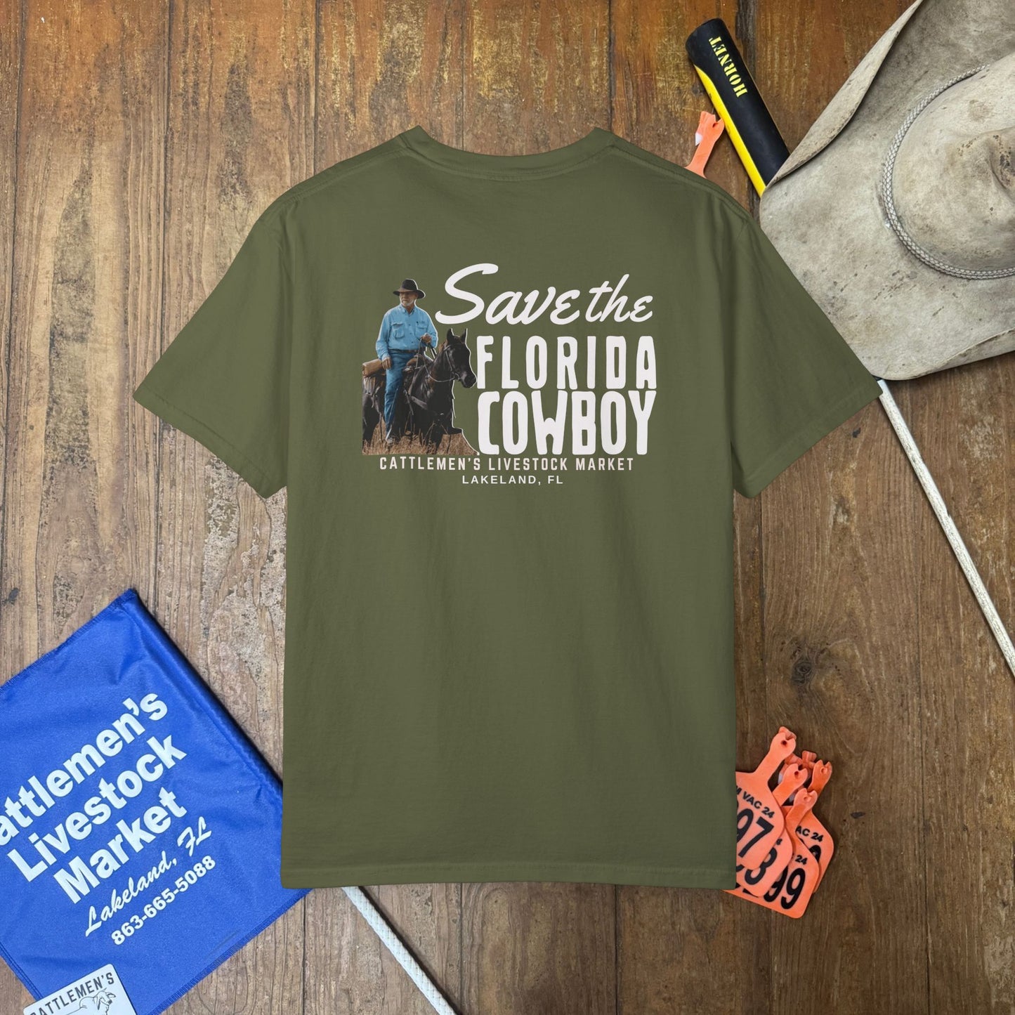 An olive green t-shirt with a graphic on the back reading 'Save the FLORIDA COWBOY' is displayed in a flat-lay arrangement on a wooden background, surrounded by cowboy-themed props including a hat, a blue sign, and cattle tags.